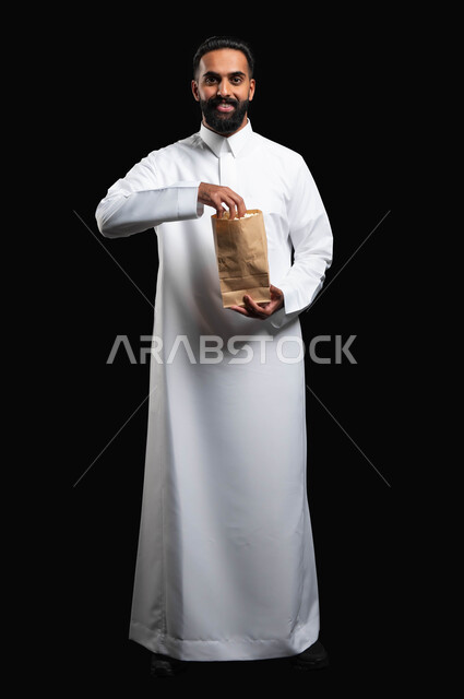 الوقوف باستقامة والنظر إلى الكاميرا بايماءات السعادة والسرور، تناول الوجبات الخفيفة، بورتريه لرجل عربي خليجي سعودي يرتدي الثوب التقليدي يحمل بيده كيس من الفوشار، الاستمتاع بمشاهدة الأفلام والمسلسلات، متابعة برامج التلفاز، قضاء وقت ممتع في المنزل، خلفية سوداء