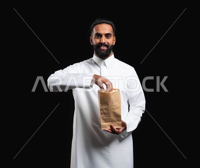 تناول الوجبات الخفيفة والسناك، انشطة ترفيهية منزلية، ايماءات السعادة والسرور، بورتريه لرجل عربي خليجي سعودي يرتدي الثوب التقليدي الأبيض يحمل بيده كيس من الفوشار، الاستمتاع بمشاهدة الأفلام والمسلسلات، متابعة برامج التلفاز، قضاء وقت ممتع في المنزل، خلفية سوداء