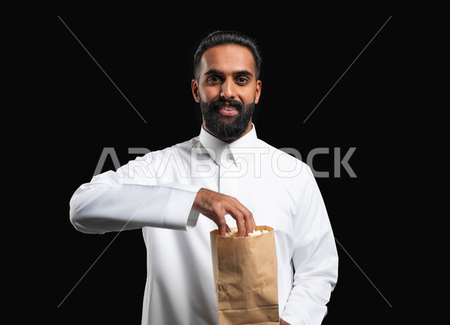 النظر إلى الكاميرا بايماءات السعادة والسرور، تناول الوجبات الخفيفة والسناك، بورتريه مقرب لرجل عربي خليجي سعودي يرتدي الثوب التقليدي الأبيض يحمل بيده كيس من الفوشار، الاستمتاع بمشاهدة الأفلام والمسلسلات، متابعة برامج التلفاز، قضاء وقت ممتع في المنزل، خلفية سوداء