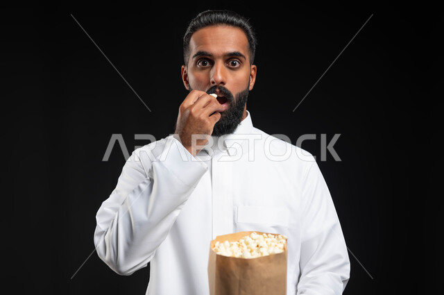 ايماءات التعجب والدهشة، بورتريه مقرب لرجل عربي خليجي سعودي يرتدي الثوب التقليدي الأبيض يحمل بيده كيس من الفوشار، الاستمتاع بمشاهدة الأفلام والمسلسلات، متابعة برامج التلفاز، تناول الوجبات الخفيفة والسناك، قضاء وقت ممتع في المنزل، انشطة ترفيهية منزلية، خلفية سوداء