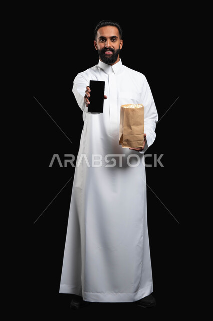 اظهار شاشة الموبايل، قضاء وقت ممتع في المنزل، الاستمتاع بمشاهدة الأفلام والمسلسلات، بورتريه لرجل عربي خليجي سعودي يرتدي الثوب التقليدي الأبيض يحمل بيده كيس من الفوشار وهاتف محمول، تناول الوجبات الخفيفة والسناك، استخدام التكنولوجيا الحديثة، خلفية سوداء