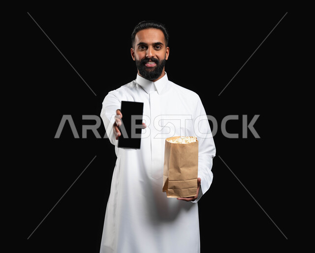 استخدام التكنولوجيا الحديثة، اظهار شاشة الموبايل، قضاء وقت ممتع في المنزل، الاستمتاع بمشاهدة الأفلام والمسلسلات، بورتريه لرجل عربي خليجي سعودي يرتدي الثوب التقليدي الأبيض يحمل بيده كيس من الفوشار وهاتف محمول، تناول الوجبات الخفيفة والسناك، خلفية سوداء
