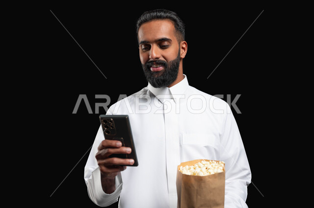 ايماءات التركيز والاندماج، قضاء وقت ممتع في المنزل، الاستمتاع بمشاهدة الأفلام والمسلسلات، بورتريه مقرب لرجل عربي خليجي سعودي يرتدي الثوب التقليدي الأبيض يحمل بيده كيس من الفوشار وهاتف محمول، تناول الوجبات الخفيفة والسناك، استخدام التكنولوجيا الحديثة، خلفية سوداء