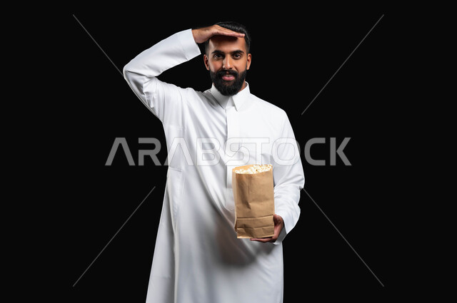 النظر إلى الكاميرا بايماءات البحث و الترقب، الاستمتاع بمشاهدة الأفلام والمسلسلات، بورتريه لرجل عربي خليجي سعودي يرتدي الثوب التقليدي الأبيض يحمل بيده كيس من الفوشار يضع يده على جبينه، تناول الوجبات الخفيفة والسناك، متابعة برامج التلفاز، خلفية سوداء