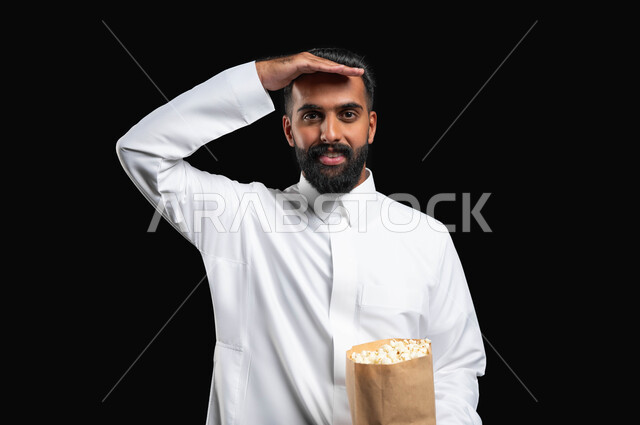 قضاء وقت ممتع في المنزل، الاستمتاع بمشاهدة الأفلام والمسلسلات، ايماءات التركيز والاندماج، بورتريه مقرب لرجل عربي خليجي سعودي يرتدي الثوب التقليدي الأبيض يحمل بيده كيس من الفوشار يضع يده على جبينه، تناول الوجبات الخفيفة والسناك، متابعة برامج التلفاز، خلفية سوداء