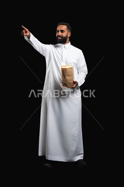 ايماءات التركيز والاندماج، بورتريه لرجل عربي خليجي سعودي يرتدي الثوب التقليدي الأبيض يحمل بيده كيس من الفوشار يشير بيده إلى شيء ما، الاستمتاع بمشاهدة الأفلام والمسلسلات، تناول الوجبات الخفيفة والسناك، متابعة برامج التلفاز، قضاء وقت ممتع في المنزل، خلفية سوداء