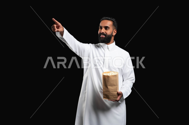 الاستمتاع بمشاهدة الأفلام والمسلسلات، ايماءات التركيز والاندماج، بورتريه لرجل عربي خليجي سعودي يرتدي الثوب التقليدي الأبيض يحمل بيده كيس من الفوشار يشير بيده إلى شيء ما، تناول الوجبات الخفيفة والسناك، متابعة برامج التلفاز، قضاء وقت ممتع في المنزل، خلفية سوداء