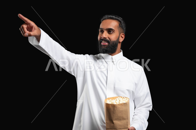 قضاء وقت ممتع في المنزل، الاستمتاع بمشاهدة الأفلام والمسلسلات، ايماءات التركيز والاندماج، بورتريه لرجل عربي خليجي سعودي يرتدي الثوب التقليدي الأبيض يحمل بيده كيس من الفوشار يشير بيده إلى شيء ما، تناول الوجبات الخفيفة والسناك، متابعة برامج التلفاز، خلفية سوداء
