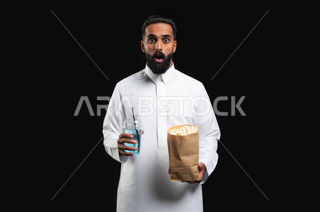الفوشار والمشروبات الباردة، انشطة ترفيهية منزلية، ايماءات التعجب والدهشة، بورتريه مقرب لرجل عربي خليجي سعودي يرتدي الثوب التقليدي الأبيض، الاستمتاع بمشاهدة الأفلام والمسلسلات، متابعة برامج التلفاز، قضاء وقت ممتع في المنزل، خلفية سوداء