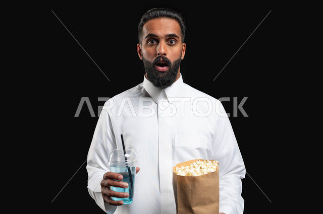 تناول الفوشار والمشروبات الباردة، انشطة ترفيهية منزلية، ايماءات التعجب والدهشة، بورتريه مقرب لرجل عربي خليجي سعودي يرتدي الثوب التقليدي الأبيض، الاستمتاع بمشاهدة الأفلام والمسلسلات، متابعة برامج التلفاز، قضاء وقت ممتع في المنزل، خلفية سوداء