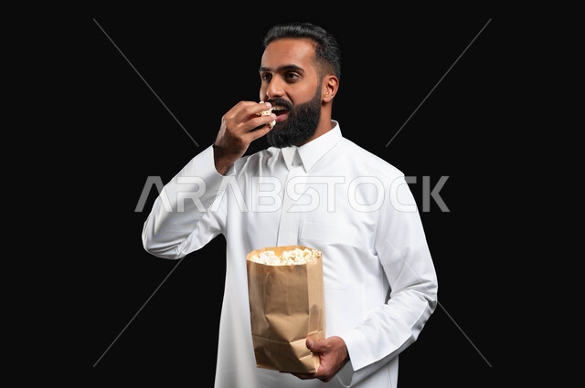 ايماءات التعجب والدهشة، بورتريه مقرب لرجل عربي خليجي سعودي يرتدي الثوب التقليدي الأبيض يحمل بيده كيس من الفوشار، الاستمتاع بمشاهدة الأفلام والمسلسلات، متابعة برامج التلفاز، تناول الوجبات الخفيفة والسناك، قضاء وقت ممتع في المنزل، انشطة ترفيهية منزلية، خلفية سوداء