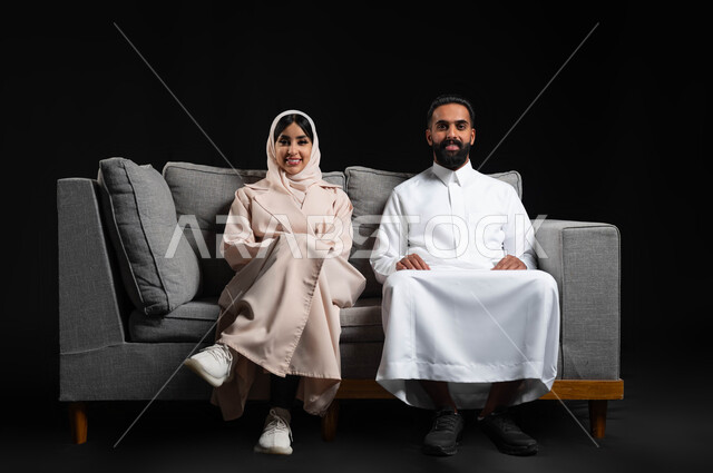 قضاء وقت ممتع في المنزل، ايماءات السعادة والسرور، متابعة المسلسلات والبرامج التفزيونية، بورتريه لزوجين عربيين خليجيين سعوديين يجلسان على كنبه مريحة يقومان بمشاهدة الافلام، أنشطة عطلة نهاية الأسبوع، صورة لكامل الجسم، خلفية سوداء