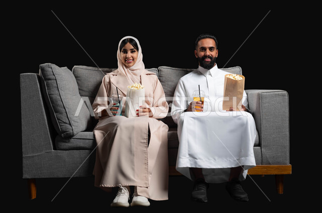 تعابير السرور والسعادة، الاستمتاع بقضاء أوقات رومانسية، تناول الفوشار والمشروبات الباردة، متابعة المسلسلات والبرامج التفزيونية، بورتريه لزوجين عربيين خليجيين سعوديين يجلسان على كنبه مريحة يقومان بمشاهدة الافلام، أنشطة عطلة نهاية الأسبوع، خلفية سوداء