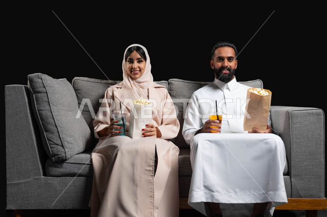 ايماءات السرور والسعادة، الاستمتاع بقضاء أوقات رومانسية، تناول الفوشار والمشروبات الباردة، متابعة المسلسلات والبرامج التفزيونية، بورتريه لزوجين عربيين خليجيين سعوديين يجلسان على كنبه مريحة يقومان بمشاهدة الافلام، أنشطة عطلة نهاية الأسبوع، خلفية سوداء