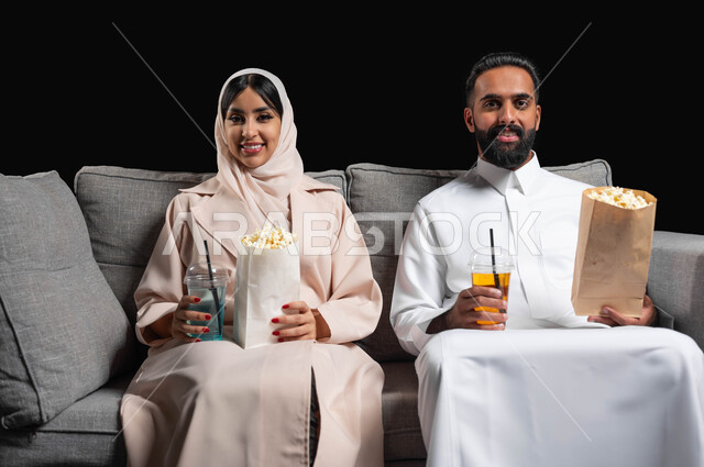 متابعة المسلسلات والبرامج التفزيونية، تعابير السرور والسعادة، الاستمتاع بقضاء أوقات رومانسية، تناول الفوشار والمشروبات الباردة، بورتريه لزوجين عربيين خليجيين سعوديين يجلسان على كنبه مريحة يقومان بمشاهدة الافلام، أنشطة عطلة نهاية الأسبوع، خلفية سوداء