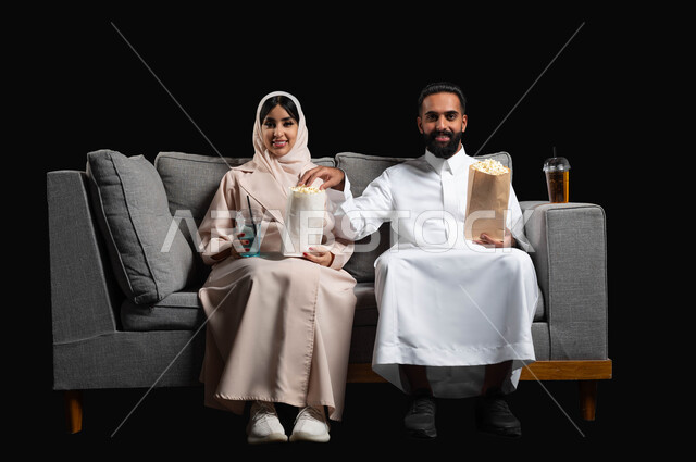مشاركة الوجبات الخفيفة مع العائلة، متابعة المسلسلات والبرامج التفزيونية، الاستمتاع بقضاء أوقات رومانسية، تناول الفوشار والمشروبات الباردة، بورتريه لزوجين عربيين خليجيين سعوديين يجلسان على كنبه مريحة يقومان بمشاهدة الافلام، أنشطة عطلة نهاية الأسبوع، خلفية سوداء