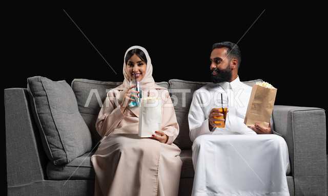 الاستمتاع بقضاء أوقات رومانسية، تناول الفوشار والمشروبات الباردة، متابعة المسلسلات والبرامج التفزيونية، ايماءات الحماس والسعادة، بورتريه لزوجين عربيين خليجيين سعوديين يجلسان على كنبه مريحة يقومان بمشاهدة الافلام، أنشطة عطلة نهاية الأسبوع، خلفية سوداء