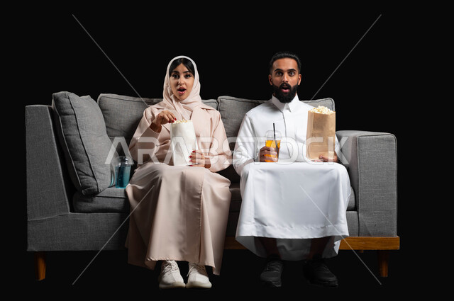 Gestures of enthusiasm and happiness, enjoying romantic times, eating popcorn and cold drinks, watching TV series and programs, portrait of a Saudi Gulf Arab couple sitting on a comfortable sofa watching movies, weekend activities, black background