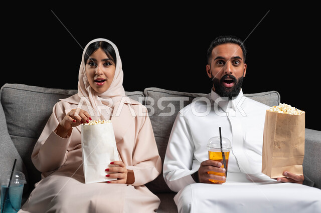 تعابير الحماس والسعادة، الاستمتاع بقضاء أوقات رومانسية، تناول الفوشار والمشروبات الباردة، متابعة المسلسلات والبرامج التفزيونية، بورتريه لزوجين عربيين خليجيين سعوديين يجلسان على كنبه مريحة يقومان بمشاهدة الافلام، أنشطة عطلة نهاية الأسبوع، خلفية سوداء