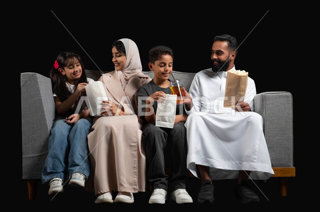 الاستمتاع بتناول الفوشار والمشروبات الباردة، أنشطة عطلة نهاية الاسبوع الترفيهية، أجواء عائلية سعيدة، مشاركة الوجبات الخفيفة مع العائلة، بورتريه أسرة عربية خليجية سعودية يجلسون على كنبة مريحة يقومون بمتابعة التلفاز، مشاهدة الافلام والمسلسلات، خلفية سوداء