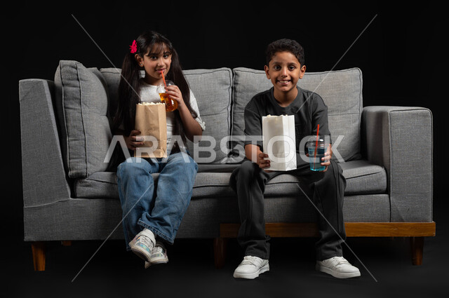 تناول الفوشار والمشروبات الباردة، ايماءات السعادة والسرور، بورتريه لأخ وأخت عربيين خليجيين سعوديين يرتديان الزي الكاجوال يجلسان على كنبة مريحة يقومان بقضاء وقت ممتع بالمنزل، الاستمتاع بمشاهدة الأفلام، برامج ترفيهية للأطفال، أنشطة عطلة نهاية الاسبوع، خلفية سوداء