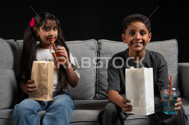 برامج ترفيهية للأطفال، الاستمتاع بمشاهدة الأفلام، أنشطة عطلة نهاية الاسبوع، قضاء وقت ممتع مع الأخوة، بورتريه لطفلة عربية خليجية سعودية تجلس على كنبة مريحة تقوم بمتابعة التلفاز مع شقيقها، تناول الفوشار والمشروبات الباردة، خلفية سوداء