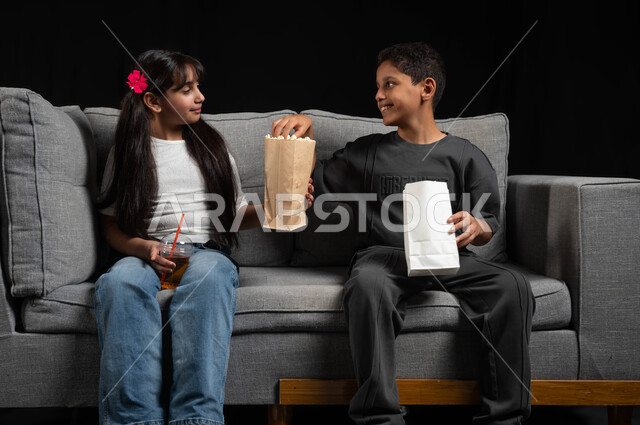 مفهوم المشاركة وقضاء وقت ممتع مع الأخوة، تناول الفوشار والمشروبات الباردة، أنشطة عطلة نهاية الاسبوع، بورتريه لأخ وأخت عربيين خليجيين سعوديين يرتديان الزي الكاجوال يجلسان على الكنبة يقومان بمشاهدة الأفلام والبرامج الترفيهية للأطفال، خلفية سوداء
