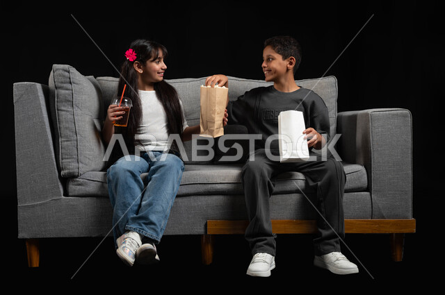 تناول الفوشار والمشروبات الباردة، ايماءات السعادة والسرور، بورتريه لأخ وأخت عربيين خليجيين سعوديين يرتديان الزي الكاجوال يجلسان على الكنبة يقومان بقضاء وقت ممتع بالمنزل، الاستمتاع بمشاهدة الأفلام والبرامج الترفيهية للأطفال، أنشطة عطلة نهاية الاسبوع، خلفية سوداء