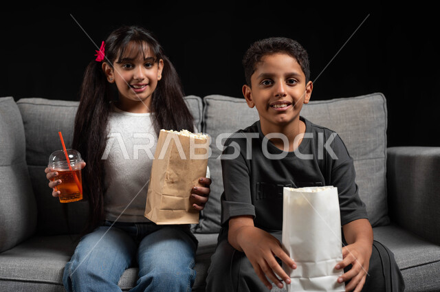 مفهوم الحب بين الأخوة، تناول الفوشار والمشروبات الباردة، بورتريه لأخ وأخت عربيين خليجيين سعوديين يرتديان الزي الكاجوال يجلسان على الكنبة يقومان بقضاء وقت ممتع بالمنزل، الاستمتاع بمشاهدة الأفلام والبرامج الترفيهية للأطفال، أنشطة عطلة نهاية الاسبوع، خلفية سوداء