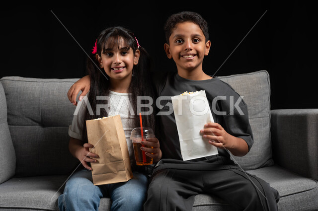 مفهوم الحب بين الأخوة، تناول الفوشار والمشروبات الباردة، بورتريه لأخ وأخت عربيين خليجيين سعوديين يرتديان الزي الكاجوال يجلسان على الكنبة يقومان بقضاء وقت ممتع بالمنزل، الاستمتاع بمشاهدة الأفلام والبرامج الترفيهية للأطفال، أنشطة عطلة نهاية الاسبوع، خلفية سوداء