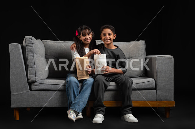 Looking at the camera with happy expressions, spending quality time with brothers, weekend activities, portrait of a Saudi Gulf Arab brother and sister wearing casual clothes sitting on the sofa eating popcorn and watching movies and entertainment programs for children, black background
