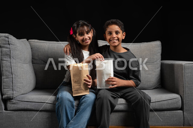 النظر إلى الكاميرا بتعابير السعادة، قضاء وقت ممتع مع الأخوة، أنشطة عطلة نهاية الاسبوع، بورتريه لأخ وأخت عربيين خليجيين سعوديين يرتديان الزي الكاجوال يجلسان على الكنبة يقومان بتناول الفوشار ومشاهدة الأفلام والبرامج الترفيهية للأطفال، خلفية سوداء