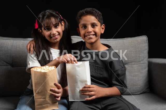 الترفيه عن النفس في عطلة نهاية الأسبوع، النظر إلى الكاميرا بتعابير السعادة، قضاء وقت ممتع مع الأخوة، بورتريه لأخ وأخت عربيين خليجيين سعوديين يرتديان الزي الكاجوال يجلسان على الكنبة يقومان بتناول الفوشار ومشاهدة الأفلام والبرامج الترفيهية للأطفال، خلفية سوداء