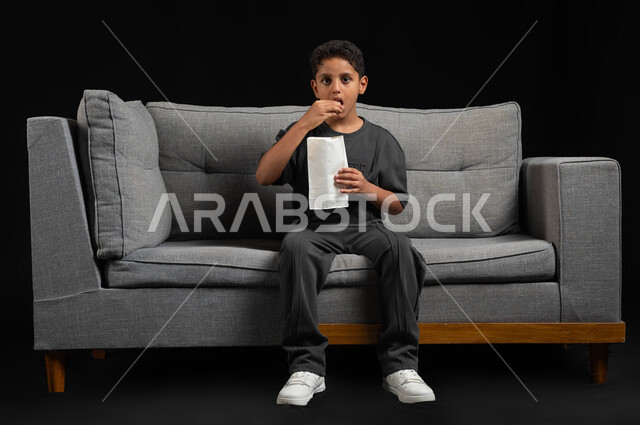 ايماءات وتعابير الاندماج والسرور، قضاء أوقات ممتعة ومسلية في المنزل، الاستمتاع بمشاهدة الأفلام والمسلسلات، بورتريه مقرب لولد عربي خليجي سعودي مبتسم يرتدي الزي الكاجوال يجلس على اريكة رمادية يقوم بتناول الفوشار، خلفية سوداء