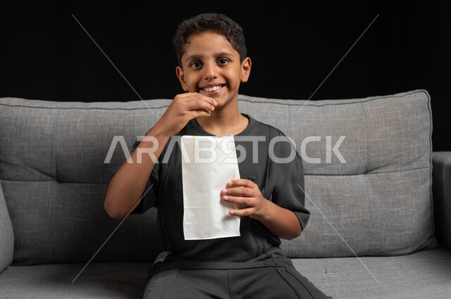 الترفيه عن النفس، ايماءات وتعابير الاندماج والسرور، قضاء أوقات ممتعة ومسلية في المنزل، الاستمتاع بمشاهدة الأفلام والمسلسلات، بورتريه مقرب لولد عربي خليجي سعودي مبتسم يرتدي الزي الكاجوال يجلس على اريكة رمادية يقوم بتناول الفوشار، خلفية سوداء