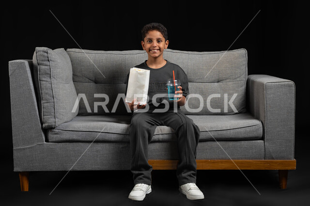Eating popcorn and cold drinks and enjoying watching movies and series, portrait of a smiling Saudi Gulf Arab boy wearing a casual outfit sitting on a gray sofa spending fun and entertaining times at home, gestures and expressions of integration and pleasure, black background