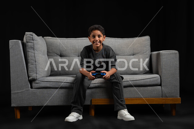 قضاء اوقات ممتعة باللعب في المنزل، استخدام التقنيات الحديثة للتسلية والترفيه، بورتريه لولد عربي خليجي سعودي مبتسم يجلس على اريكة رمادية يحمل عصا التحكم يقوم باللعب في ألعاب الفيديو، تعابير وايماءات الاندماج والتركيز، خلفية سوداء