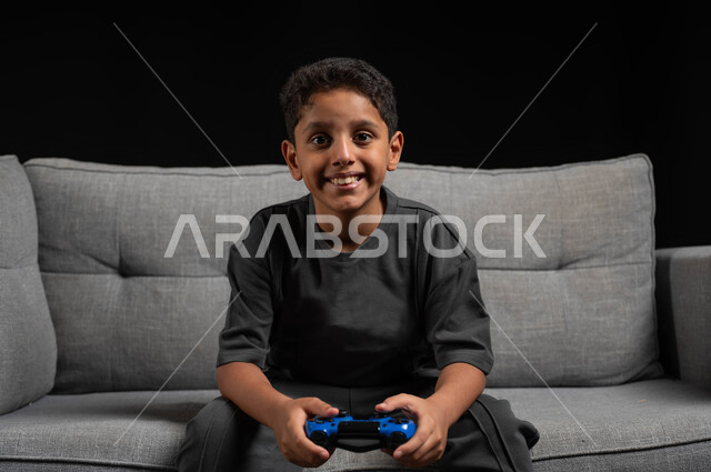 Using modern technologies for fun and entertainment, a close-up portrait of a smiling Saudi Gulf Arab boy sitting on a gray sofa, holding a joystick, playing video games, expressions and gestures of integration and concentration, spending enjoyable times playing, black background