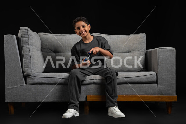 قضاء اوقات ممتعة باللعب في المنزل، استخدام التقنيات الحديثة للتسلية والترفيه، بورتريه لولد عربي خليجي سعودي مبتسم يجلس على اريكة رمادية يحمل عصا التحكم يقوم باللعب في ألعاب الفيديو، تعابير وايماءات الاندماج والتركيز، خلفية سوداء