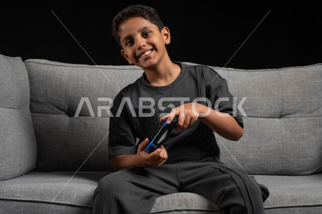 استخدام التقنيات الحديثة للتسلية والترفيه، بورتريه مقرب لولد عربي خليجي سعودي مبتسم يجلس على اريكة رمادية يحمل عصا التحكم يقوم باللعب في ألعاب الفيديو، تعابير وايماءات الاندماج والتركيز، قضاء اوقات ممتعة باللعب، خلفية سوداء