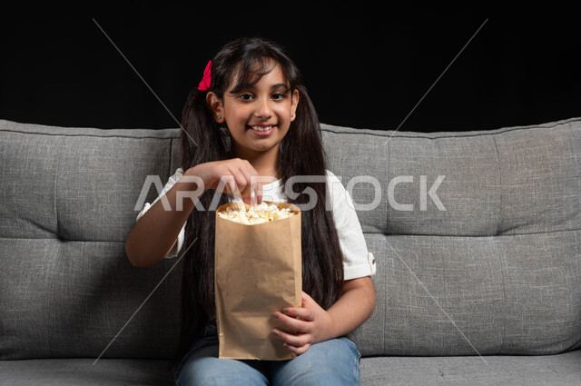 الاستمتاع في عطلة نهاية الإسبوع، قضاء أوقات ممتعة ومسلية في المنزل، متابعة الأفلام والسينما والمسلسلات، بورتريه لفتاة عريية خليجية سعودية مبتسمة تجلس على كنبة رمادية تقوم بمشاهدة الأفلام وتناول الفوشار بتعابير وايماءات الاندماج والتركيز، خلفية سوداء