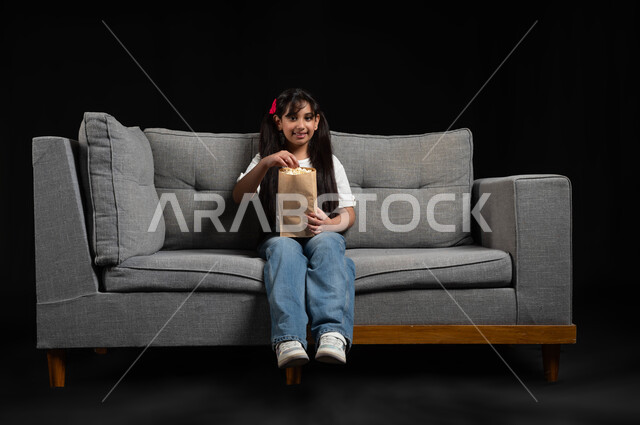 قضاء أوقات ممتعة ومسلية في المنزل، الاستمتاع في عطلة نهاية الإسبوع، متابعة الأفلام والسينما والمسلسلات، بورتريه لفتاة عريية خليجية سعودية مبتسمة تجلس على كنبة رمادية تقوم بمشاهدة الأفلام وتناول الفوشار بتعابير وايماءات الاندماج والتركيز، خلفية سوداء