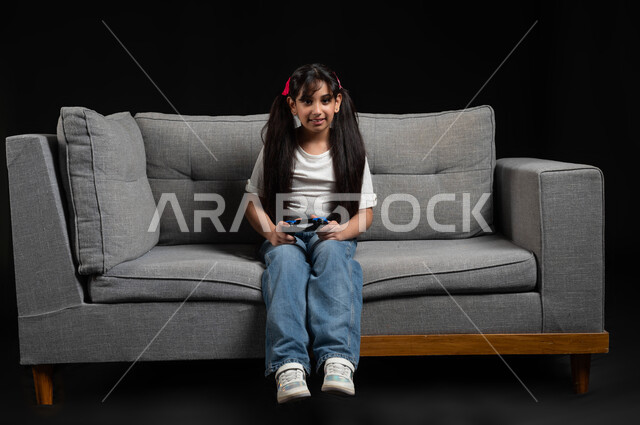 قضاء اوقات ممتعة باللعب في المنزل، استخدام التقنيات الحديثة للتسلية والترفيه، بورتريه لبنت عربية خليجية سعودية مبتسمة ترتدي الزي الكاجوال تجلس على اريكة رمادية تقوم باللعب في ألعاب الفيديو بتعابير وايماءات الاندماج والتركيز، خلفية سوداء