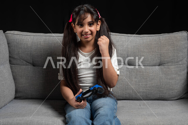 إيماءات الفوز والانتصار، قضاء اوقات ممتعة باللعب في المنزل، استخدام التقنيات الحديثة للتسلية والترفيه، بورتريه لبنت عربية خليجية سعودية مبتسمة ترتدي الزي الكاجوال تجلس على اريكة رمادية تقوم باللعب في ألعاب الفيديو بتعابير الاندماج والتركيز، خلفية سوداء