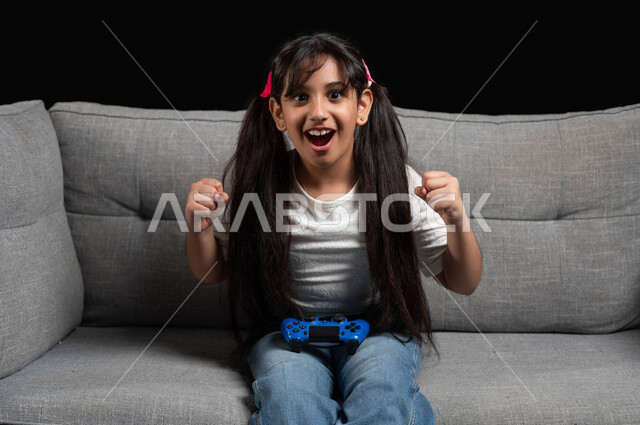 استخدام التقنيات الحديثة للتسلية والترفيه، إيماءات الفوز والانتصار، قضاء اوقات ممتعة باللعب في المنزل، بورتريه لبنت عربية خليجية سعودية مبتسمة ترتدي الزي الكاجوال تجلس على اريكة رمادية تقوم باللعب في ألعاب الفيديو بتعابير السعادة والسرور، خلفية سوداء