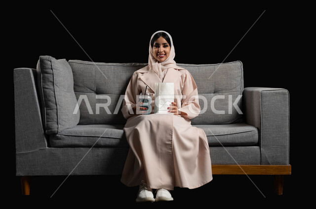 تناول الفوشار والمشروبات الباردة والاستمتاع بمشاهدة الأفلام والمسلسلات، بورتريه لإمرأة عربية خليجية سعودية مبتسمة ترتدي الحجاب والعباءة تجلس على اريكة رمادية تقضي أوقات ممتعة ومسلية في المنزل، ايماءات وتعابير الاندماج والسرور، خلفية سوداء