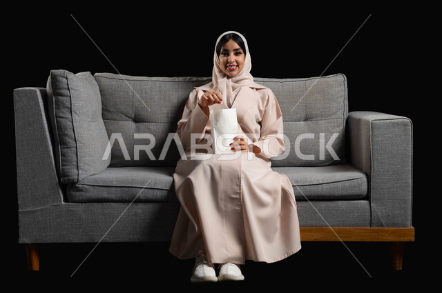 قضاء أوقات ممتعة ومسلية في المنزل، ايماءات وتعابير الاندماج والسرور، الاستمتاع بمشاهدة الأفلام والمسلسلات، بورتريه لشابة عربية خليجية سعودية مبتسمة ترتدي الحجاب والعباءة تجلس على اريكة رمادية تقوم بمشاهدة التلفاز وتناول الفوشار، خلفية سوداء