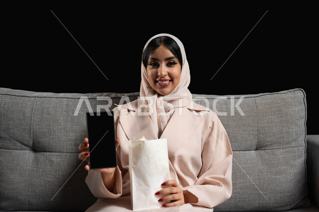 Gestures of happiness and pleasure, displaying a blank black mobile phone screen, spending enjoyable times watching movies and series, using modern technical devices, a portrait of a smiling Saudi Arabian Gulf woman wearing a hijab sitting on a gray sofa holding a bag of popcorn in her hand, black background