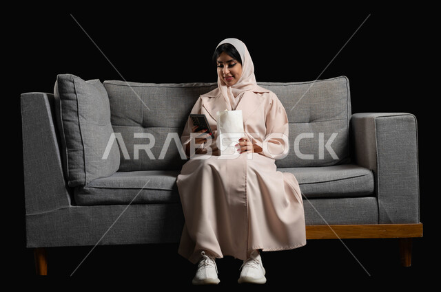 الاستمتاع بمشاهدة الأفلام والمسلسلات في السينما، استخدام الأجهزة التكنولوجية الحديثة، بورتريه لشابة عربية سعودية خليجية مبتسمة ترتدي الحجاب والعباءة تجلس على كنبة رمادية تحمل بيدها كيس الفوشار والهاتف المحمول، ايماءات الاندماج والسرور، خلفية سوداء
