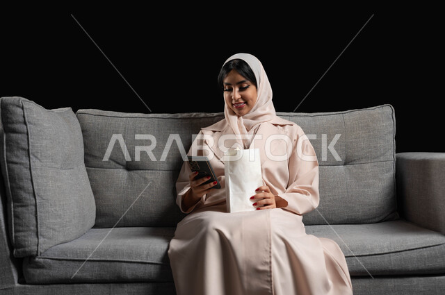 الاستمتاع بمشاهدة الأفلام والمسلسلات في السينما، استخدام الأجهزة التكنولوجية الحديثة، بورتريه لشابة عربية سعودية خليجية مبتسمة ترتدي الحجاب والعباءة تجلس على كنبة رمادية تحمل بيدها كيس الفوشار والهاتف المحمول، ايماءات الاندماج والسرور، خلفية سوداء